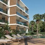 Modern residential building with glass balconies beside a pool area and tropical plants