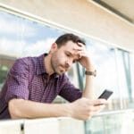 Man leaning on a railing outside looking at his smartphone with a worried expression and hand on his forehead