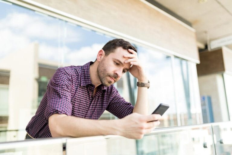 Man leaning on a railing outside looking at his smartphone with a worried expression and hand on his forehead