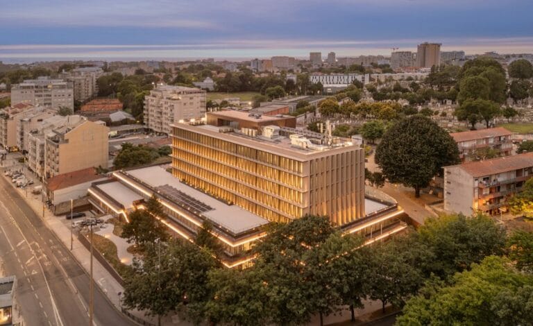 Aerial view of a modern multi story building with warm exterior lighting at dusk surrounded by trees and a city block of smaller buildings