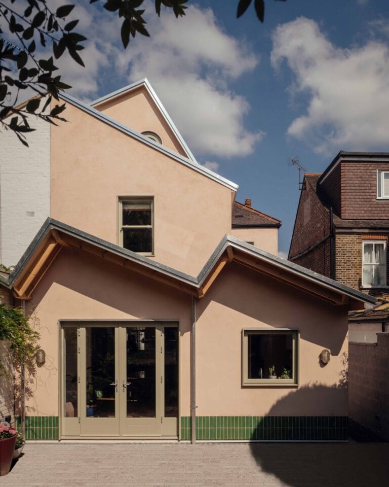 Front of a peach colored house with large glass doors and a green tiled base under a blue sky with clouds 2 story roofline nearby