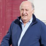 Older man in a blue jacket walking outdoors past a red door smiling slightly in daylight