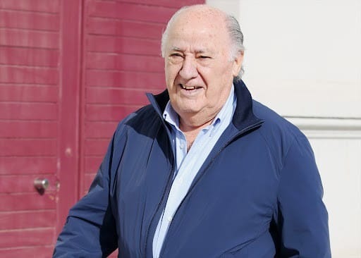 Older man in a blue jacket walking outdoors past a red door smiling slightly in daylight