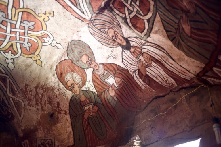 Colorful wall fresco showing several men in striped robes with folded hands in prayer next to a decorative knotwork design
