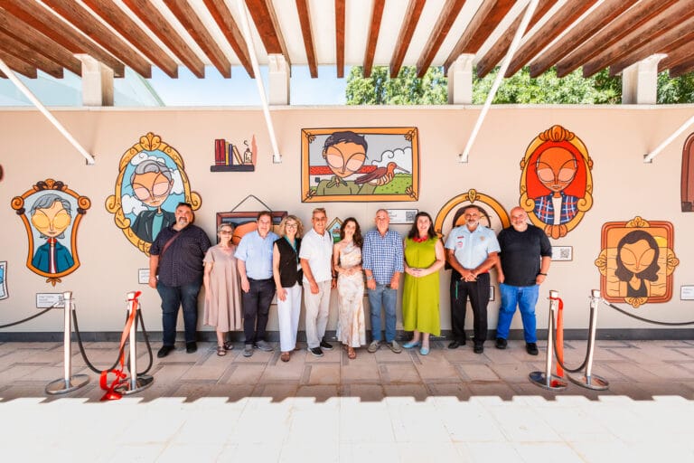 A group of twelve adults standing side by side in front of colorful cartoon portraits on a wall at an outdoor event