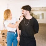 Smiling woman touches a mans shoulder as they hold hands in a bright room with unpacked boxes nearby