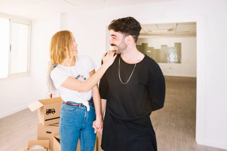 Smiling woman touches a mans shoulder as they hold hands in a bright room with unpacked boxes nearby