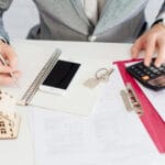 Person at a desk writing in a grid notebook with a smartphone keys and a house model nearby on a white table with documents and a calculator nearby