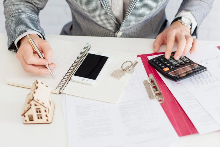 Person at a desk writing in a grid notebook with a smartphone keys and a house model nearby on a white table with documents and a calculator nearby