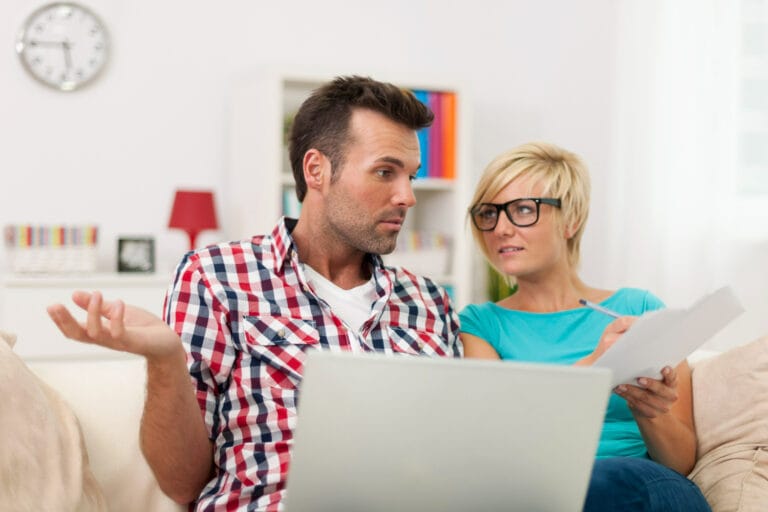 Couple on a couch looking at papers together while a laptop rests on the mans lap in a bright living room