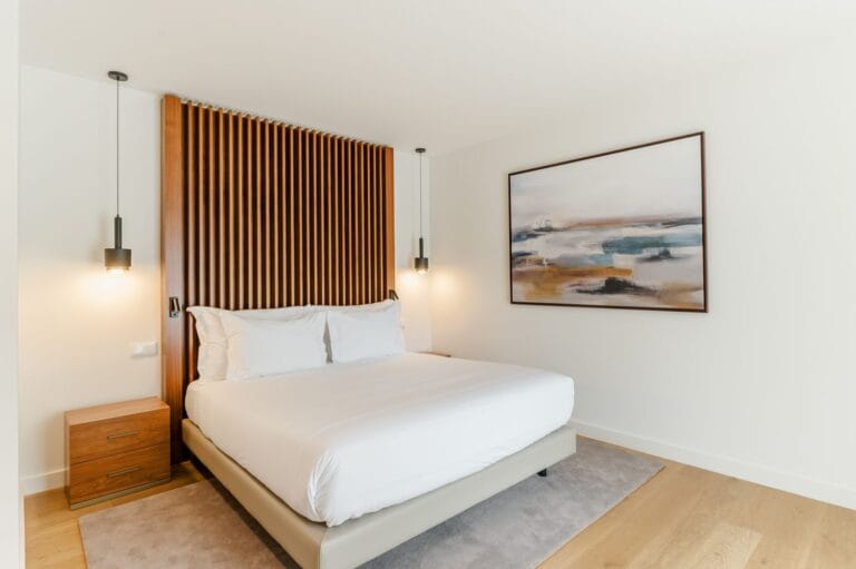 Modern bedroom featuring a white bed with a tall wooden slatted headboard pendant lights and a framed abstract painting on the wall