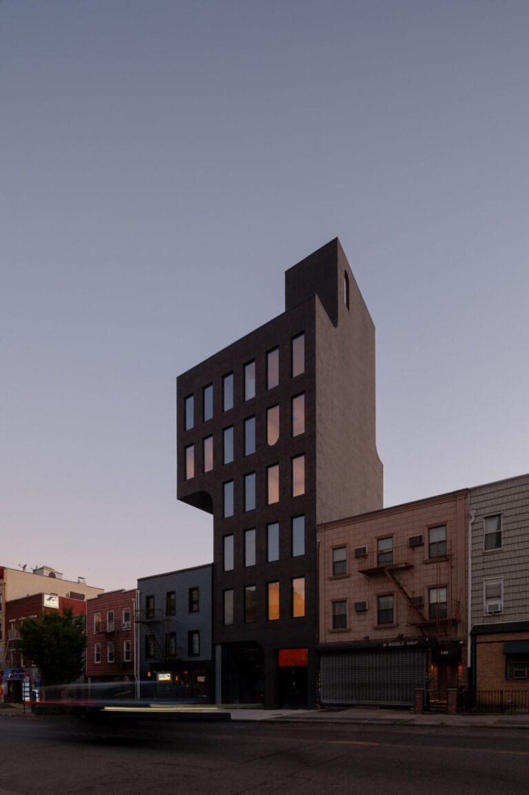 Tall dark brick building with a stepped top and evenly spaced rectangular windows against a dusky sky on a city street edge