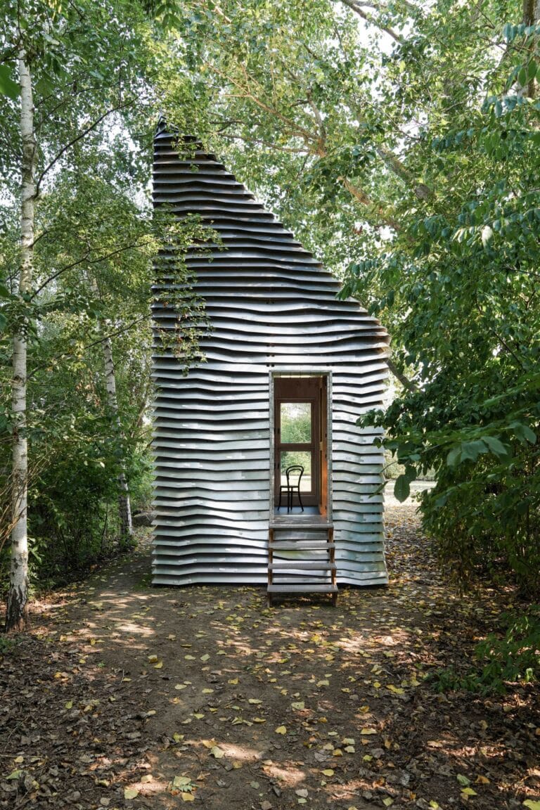 A small sculptural hut with stacked horizontal slats forming a tapered cave like shape among trees a doorway reveals a chair inside