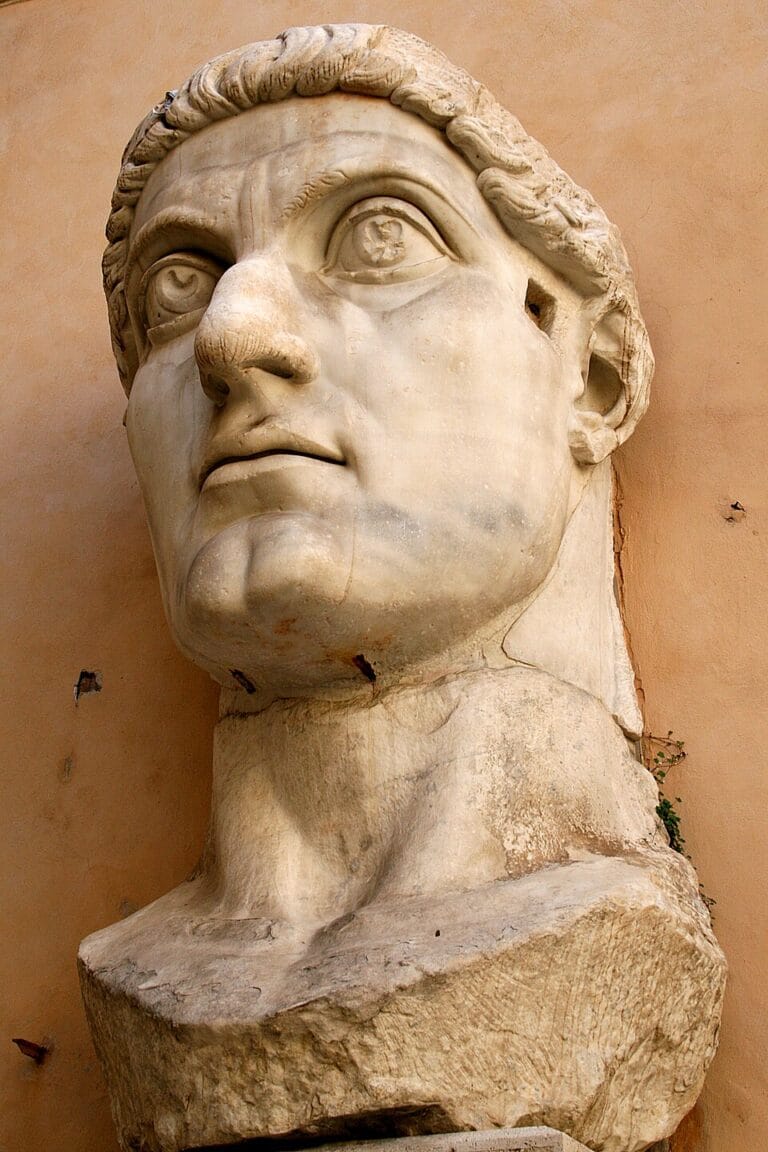 Marble bust of a young man with curly hair gazing upward on a weathered pedestal against a peach wall