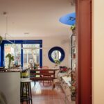 Bright kitchen and dining area with blue-framed windows and a round blue window, plants on shelves and a wooden table in the center. A view of the backyard is visible through the glass doors.