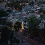 Evening view over a densely built hillside neighborhood with illuminated windows and streetlights among modern rooftops and trees