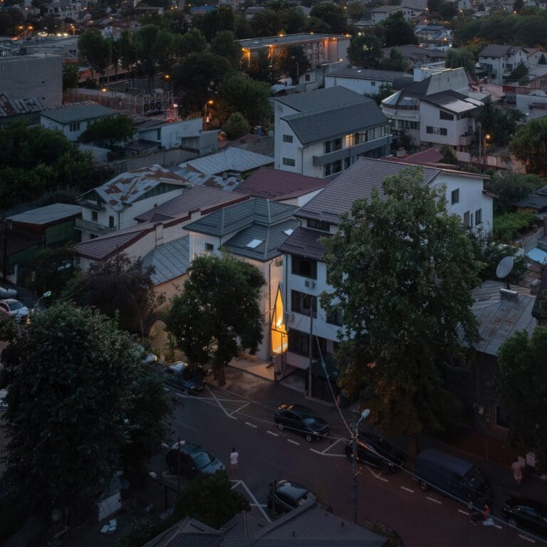 Evening view over a densely built hillside neighborhood with illuminated windows and streetlights among modern rooftops and trees