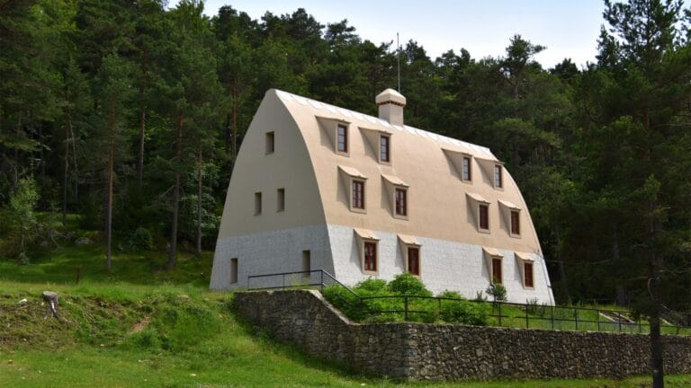 Beige curved roof house nestled in a dense pine forest with a stone retaining wall in the foreground