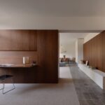 Wood paneled home office with a built in desk and a metal chair open to a living area in the background