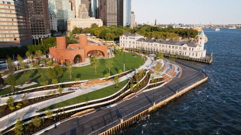 Aerial view of a waterfront park with a white historic riverside building and a red brick pavilion skyscrapers behind