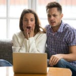 A surprised woman and a worried man sit on a couch looking at a laptop on a coffee table between them