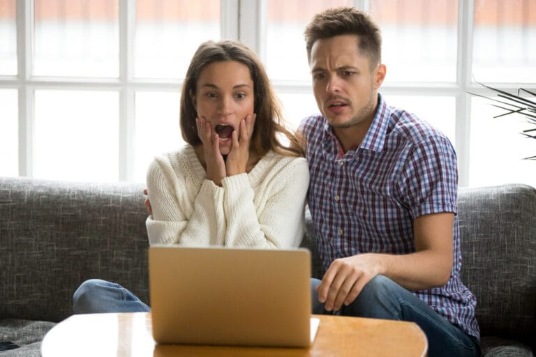 A surprised woman and a worried man sit on a couch looking at a laptop on a coffee table between them