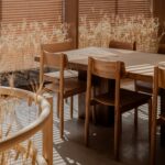 Wooden dining table and chairs in a sunlit room with vertical blinds and dried ornamental grasses by the window
