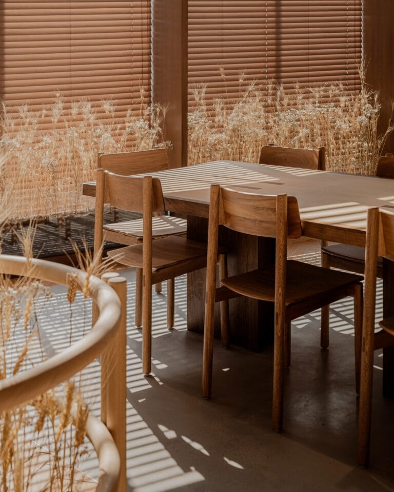Wooden dining table and chairs in a sunlit room with vertical blinds and dried ornamental grasses by the window