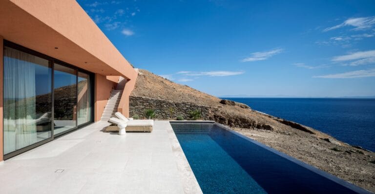 Modern peach colored house with glass walls white lounge chairs and an infinity pool overlooking a rocky hillside and the blue sea