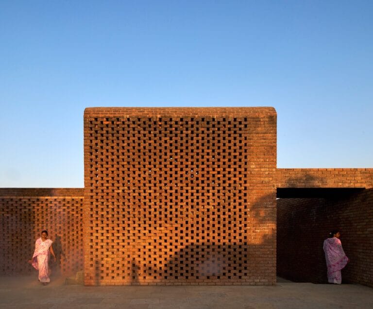 Brick lattice brick wall beneath a clear blue sky two women in pink sarees stand at ground level on opposite sides of the wall