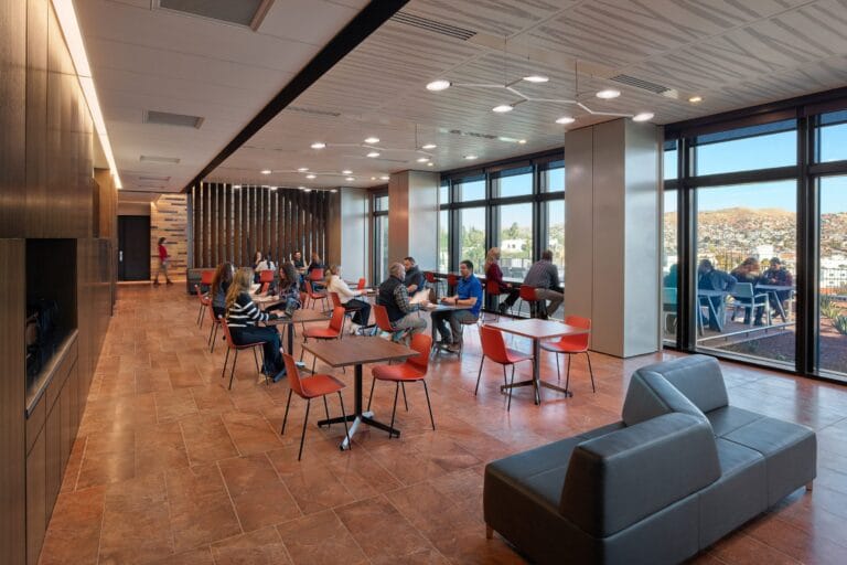 Open lounge area with red chairs and dark wood tables people sit and chat near large windows overlooking a city hillside landscape