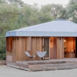 Wooden cabin with a metal roof on a raised deck two white butterfly chairs on the porch forested trees in the background