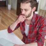 Man in a red plaid shirt sits at a white desk hand on chin looking thoughtful in a home office with a laptop and smartphone nearby