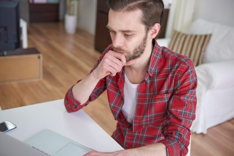 Man in a red plaid shirt sits at a white desk hand on chin looking thoughtful in a home office with a laptop and smartphone nearby