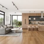 Open concept living room and kitchen with a grey sectional round coffee table marble island wooden cabinetry pendant lights and large windows showing greenery