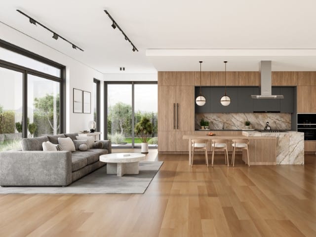 Open concept living room and kitchen with a grey sectional round coffee table marble island wooden cabinetry pendant lights and large windows showing greenery