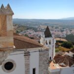 View from a stone fortress overlooking a white walled village with red tiled roofs a conical topped church tower and fields stretching to distant hills under a blue sky