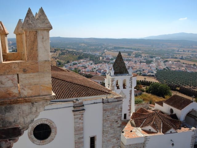 View from a stone fortress overlooking a white walled village with red tiled roofs a conical topped church tower and fields stretching to distant hills under a blue sky