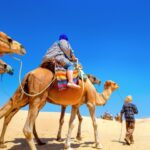 Person riding a camel in a desert, with a guide and more camels in the background under a bright blue sky.