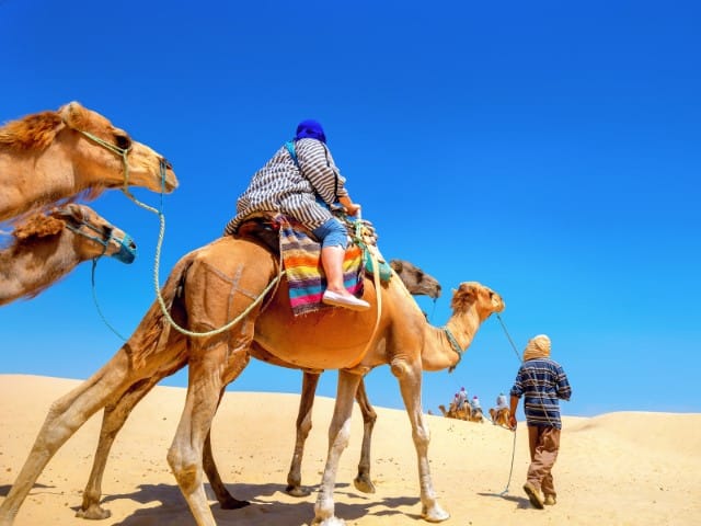 Person riding a camel in a desert with a guide and more camels in the background under a bright blue sky