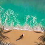 Aerial view of a person sunbathing on a sandy beach with turquoise waves washing ashore