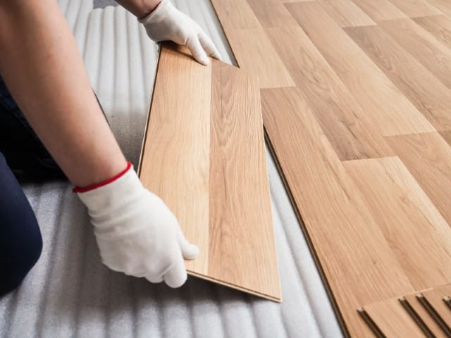 Worker wearing gloves fits a wooden floor plank into place during installation on a foam underlayment