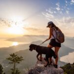 Hiker with backpack and a leashed dog standing on a rocky summit at sunset overlooking a valley and water below