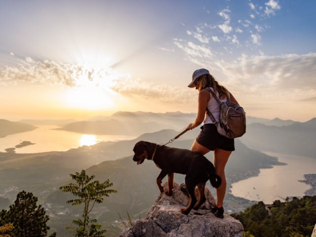 Hiker with backpack and a leashed dog standing on a rocky summit at sunset overlooking a valley and water below