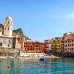 Colorful coastal town with pastel houses along a calm harbor a white bell tower on the left and small boats on the water
