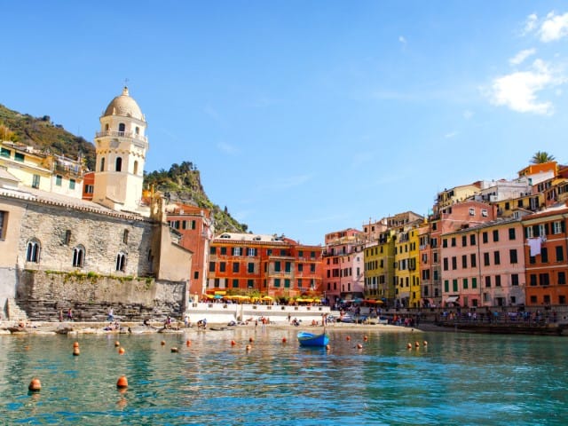 Colorful coastal town with pastel houses along a calm harbor a white bell tower on the left and small boats on the water