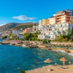 Sunny Mediterranean coastal town with white apartment buildings along a rocky shoreline and a sandy beach with straw umbrellas by turquoise water