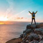 Hiker on rocky cliff with backpack raising arms at sunset over the ocean