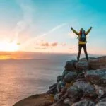 Hiker on rocky cliff with backpack raising arms at sunset over the ocean