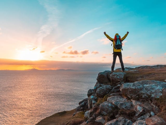 Hiker on rocky cliff with backpack raising arms at sunset over the ocean
